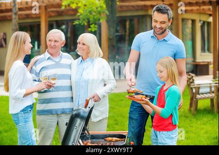 Passare un bel po' di tempo con la famiglia. Famiglia felice di cinque persone che barbecue carne alla griglia sul cortile posteriore Foto Stock