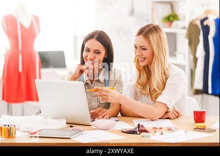 Alla ricerca delle nuove tendenze della moda. Due giovani donne gioiose che lavorano insieme mentre sono sedute alla scrivania nel loro laboratorio di moda Foto Stock