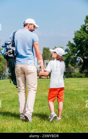 Giocatori di golf sicuri di sé. Vista posteriore del giovane e del figlio che si tengono per mano e si guardano mentre cammina sul campo da golf Foto Stock