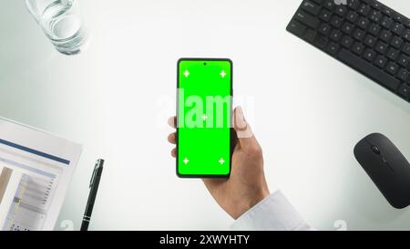 Vista dall'alto verso il basso di una persona che detiene uno smartphone con schermo Chromakey con occultamento verde e segnaposto Motion Tracker. Scrivania da ufficio con gadget per computer wireless, penna e documenti. Foto Stock