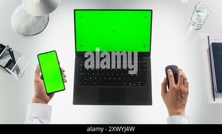 Visualizzazione dall'alto verso il basso di una persona anonima che esegue multitasking con un computer portatile e uno smartphone moderno. Entrambi i dispositivi sono dotati di schermo verde di occultamento con modelli di tracciamento del movimento. L'uomo sta usando un mouse Foto Stock