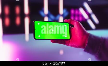 Persona del giocatore che controlla un post sul social network su smartphone con schermo Chromakey con occultamento verde. Persona che tiene il cellulare orizzontalmente in una futuristica Neon Room, streaming di contenuti di intrattenimento Foto Stock