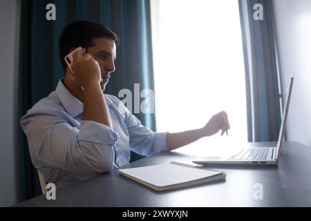 Un giovane che lavora in telelavoro da casa, risponde a una chiamata e guarda lo schermo grigio del suo notebook durante una normale giornata lavorativa Foto Stock