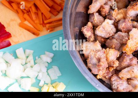 Pezzi di maiale tritati fritti in un recipiente accanto a carote e cipolle a fette vengono preparati per un'ulteriore cottura. Foto di alta qualità Foto Stock