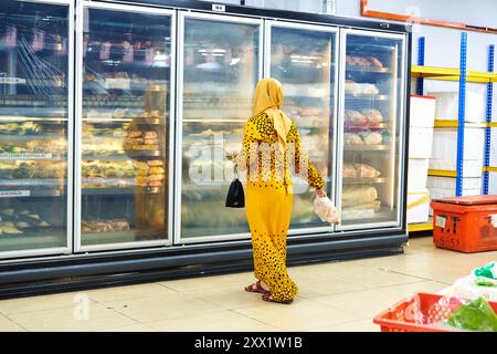 Una donna in abiti musulmani gialli sta con le spalle al negozio e sceglie il cibo vicino ai frigoriferi Foto Stock