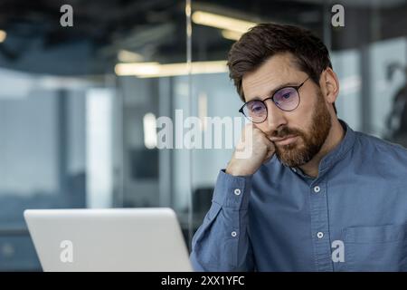 Foto ravvicinata di un giovane stanco e annoiato con una camicia blu e degli occhiali, che poggia la testa sulla mano e guarda tristemente lo schermo del notebook. Foto Stock