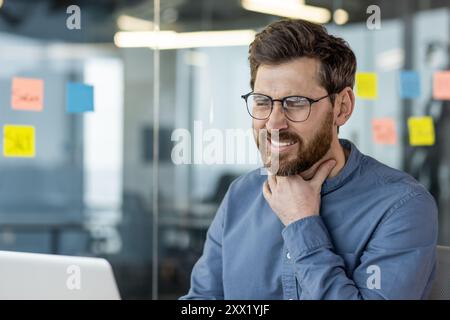 La foto ravvicinata di un giovane uomo d'affari con occhiali che è in ufficio, si siede e tiene la mano alla gola, sente un forte dolore. Foto Stock