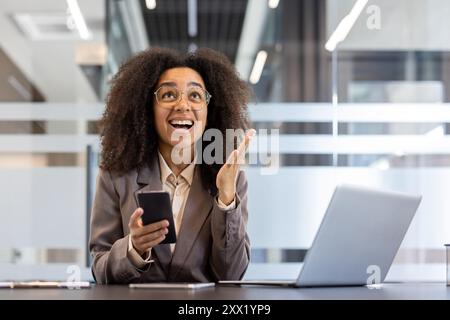 Felice giovane donna d'affari afroamericana in tuta e occhiali è felice di ricevere notizie al telefono, sorridendo alzato le mani in alto in ringraziamento. Foto Stock