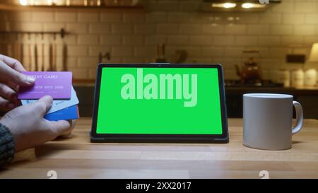 Tablet con schermo verde sul bancone della cucina. Una persona è in possesso di più carte di credito, suggerendo shopping online o gestione finanziaria in un mese Foto Stock