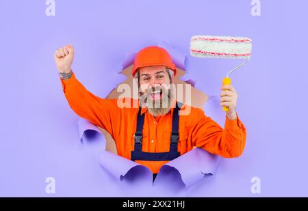 Uomo pittore con rullo per pittura che guarda attraverso il foro nella carta. Uomo con barba in abbigliamento da lavoro con rullo per pittura. Pittore o designer professionista in Foto Stock