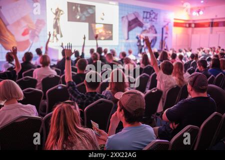 Pubblico entusiasta che ascolta la presentazione durante la conferenza. Persone che alzano la mano, interagiscono con l'altoparlante. L'atmosfera dell'evento mette in risalto la partecipazione attiva e l'interesse. Foto Stock