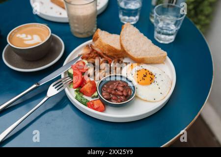 Colazione tradizionale inglese con fagioli al forno, pancetta, uova fritte, funghi pomodori, cetrioli e pane con cappuccino e un bicchiere d'acqua Foto Stock