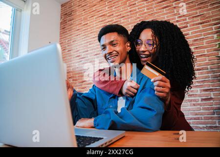 Una allegra coppia afroamericana sta usando un portatile per cercare una vendita in un negozio online. Marito e moglie entusiasti sono seduti in salotto, con una carta di credito che guarda un computer per una consegna. Foto di alta qualità Foto Stock
