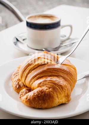 Croissant e forchetta su un piatto bianco, accompagnati da una tazza di caffè bianco in un caffè all'aperto. Foto Stock