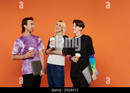 Tre giovani amici queer vestiti con un abbigliamento colorato si uniscono in uno studio con sfondo arancione. Foto Stock