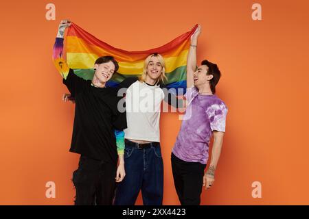 Tre giovani amici, vestiti con un abbigliamento elegante e vivace, si mettono in posa su uno sfondo arancione tenendo in mano una bandiera arcobaleno. Foto Stock