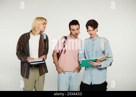 Tre giovani uomini in abbigliamento casual si stagliano insieme su uno sfondo grigio, uno legge un libro mentre gli altri lo guardano. Foto Stock