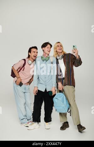 Tre giovani uomini, vestiti con un abbigliamento casual, si esibiscono in uno studio con sfondo grigio e scattano un selfie. Foto Stock