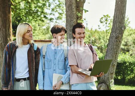 Tre giovani amici queer, vestiti in modo informale, stanno insieme in un parco, ridendo e condividendo un notebook. Foto Stock