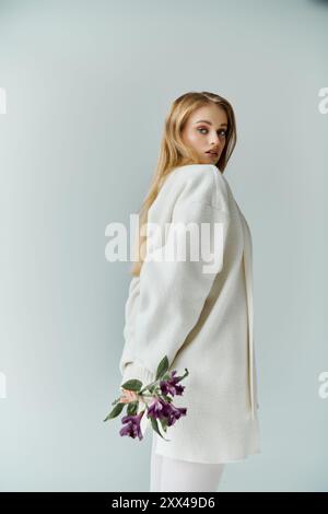 Una giovane donna con un maglione bianco tiene i fiori viola dietro la schiena, guardando sulla spalla. Foto Stock