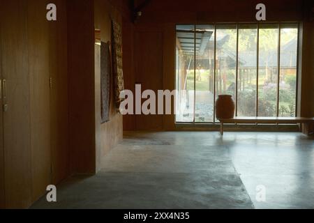 Uno spazio interno in stile giapponese con pavimenti in cemento, grandi finestre in vetro, pareti in legno e vista del giardino Foto Stock