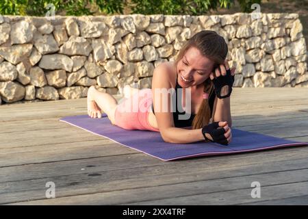 La giovane donna con i capelli rossi gode del sole, sorridendo dopo aver completato la sua routine yoga e Pilates in una giornata di sole. E' seduta sul suo tappetino, si sta godendo Foto Stock