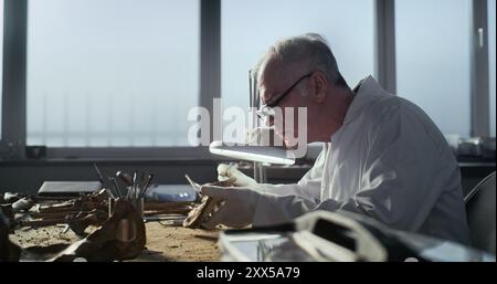 Il paleoantropologo esperto di guanti e camice da laboratorio esamina le ossa umane preistoriche sotto la lampada, utilizza strumenti professionali. Lo scienziato maschile lavora con resti fossili nel moderno laboratorio archeologico. Foto Stock