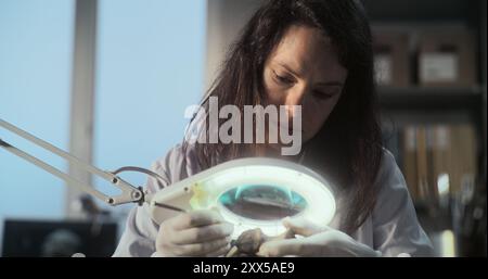 L'archeologa donna pulisce le ossa fossili utilizzando pennelli e strumenti, guarda attraverso una lampada con lente d'ingrandimento i resti di un uomo o di un animale preistorico nel laboratorio archeologico. Colleghi che lavorano in background Foto Stock