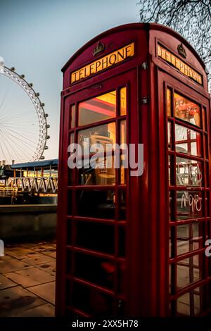 Una classica cabina telefonica rossa si trova accanto al Tamigi, con il London Eye sullo sfondo, catturando il fascino senza tempo della città al crepuscolo. Foto Stock