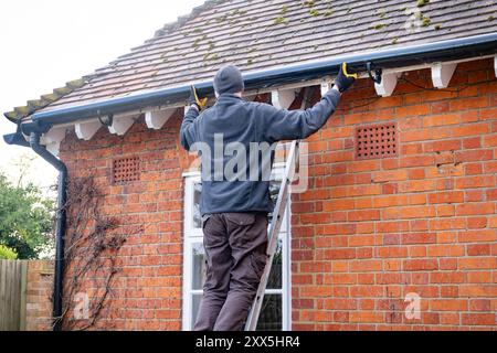 Uomo su una scala che fissa un gocciolatoio sulle grondaie di una casa del Regno Unito Foto Stock