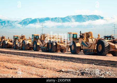 Una flotta di traslocatori di terra prepara terreni desertici aridi per lo sviluppo di nuove abitazioni. Le montagne si insinuano sullo sfondo mentre le attrezzature da costruzione si trasformano Foto Stock