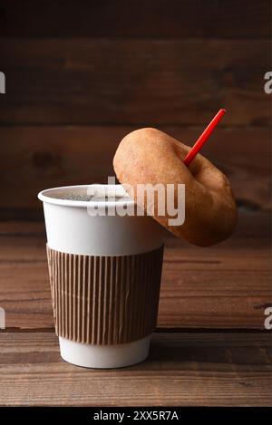 Primo piano di una tazza di carta di caffè caldo appena estratto con un cucchiaio rosso, Una ciambella semplice e vecchio stile è appesa al cucchiaio. Formato verticale con copia spa Foto Stock