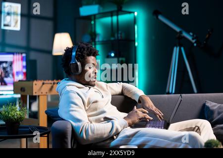 Uomo afroamericano che ascolta podcast audio mentre lavora da casa, facendo multitasking. Telelavoratore che indossa le cuffie e digita sulla tastiera del computer portatile in un appartamento illuminato al neon Foto Stock