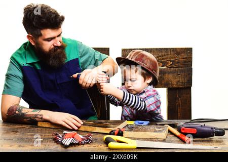 Servizio di riparazione. Uomo barbuto con un piccolo assistente. Padre che insegna al figlio piccolo a usare gli utensili manuali in officina. Il ragazzo concentrato con un cacciavite è Foto Stock