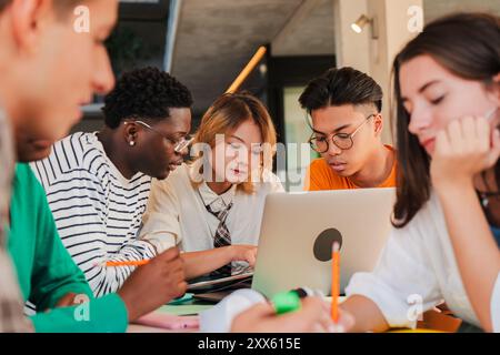 Un gruppo eterogeneo di giovani studenti collabora a un tavolo, impegnato in una sessione di studio intorno a un notebook con notebook e penne colorate, evidenziando l'importanza del lavoro di squadra nell'apprendimento Foto Stock