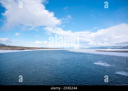 Una giornata luminosa con cielo azzurro con nuvole parziali e un grande fiume con montagne innevate e paesaggi innevati che circondano Tagish, Yukon, Canada Foto Stock