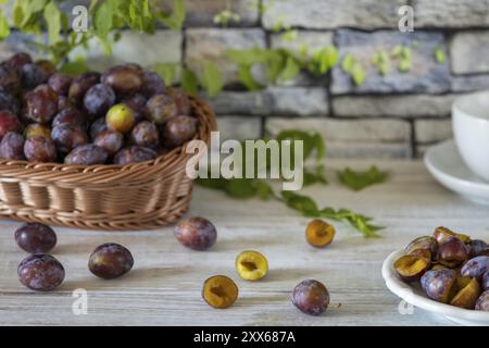 Prugne fresche intere e affettate in un cesto e su un piatto bianco, su un rustico tavolo di legno di fronte a un muro di pietra Foto Stock