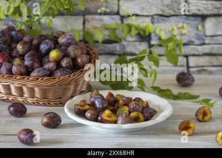 Prugne fresche intere e affettate in un cesto e su un piatto bianco, su un rustico tavolo di legno di fronte a un muro di pietra Foto Stock