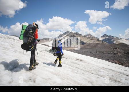 Due turisti, un uomo e una donna con zaini e ramponi ai piedi, camminano lungo il ghiacciaio sullo sfondo delle montagne del cielo Foto Stock