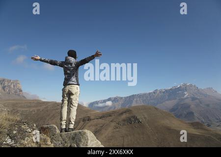 Un applauso di una giovane donna in spalla sulla cima di un'ampia cima di montagna. Libertà e vittoria sullo sfondo di montagne e blu Foto Stock