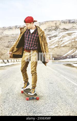 Elegante giovane felice con un berretto e pantaloni che jogger girovagano lungo una strada di montagna su un longboard, godendosi la vita Foto Stock