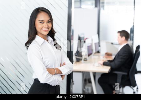 Una giovane donna d'affari sicura di sé sorridente e in piedi con le braccia incrociate in un moderno ambiente d'ufficio. Trasuda professionalità e leadership Foto Stock