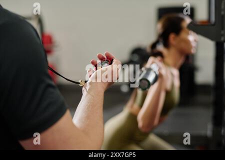Primo piano medio di un allenatore maschile irriconoscibile con la mano che tiene il cronometro rosso mentre una atleta fa gli squat su sfondo sfocato Foto Stock