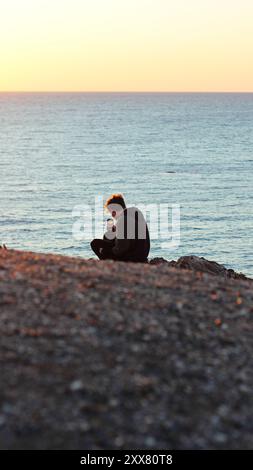 La persona si siede su una scogliera rocciosa al tramonto, catturando sul telefono Foto Stock