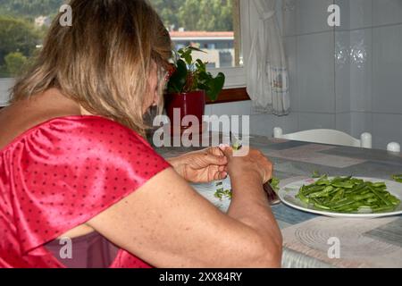 donna anziana seduta al suo tavolo della cucina, affettando con cura i fagiolini con un coltello. La scena riflette la semplicità e il comfort della vita quotidiana, wh Foto Stock