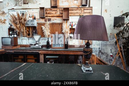 Un elegante bancone bar con una lampada da tavolo vintage con un paralume a pieghe in primo piano. Lo sfondo sfoggia un decor rustico con scaffali in legno Foto Stock