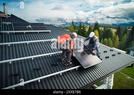 Installazione di un sistema solare in cima alla casa. Ingegneri professionisti in caschi che fissano il sistema di pannelli solari su un tetto. Due lavoratori maschi che installano un modulo solare sul tetto sullo sfondo delle montagne. Foto Stock