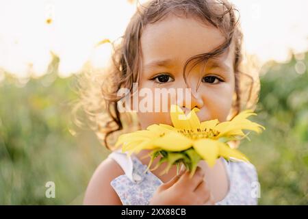 Primo piano ritratto di una bambina che tiene in mano un girasole giallo pieno fiore in un campo in campagna al tramonto con una luce calda. co Foto Stock