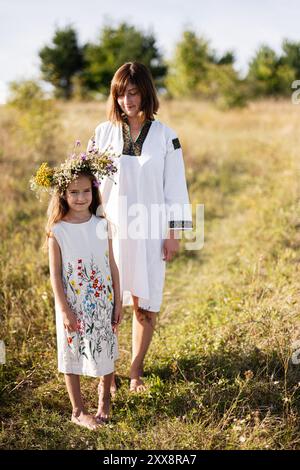 Una madre e sua figlia stanno in piedi in un prato soleggiato, entrambi indossando splendidi abiti ricamati. La ragazza indossa una corona floreale che simboleggia la natura e la t Foto Stock