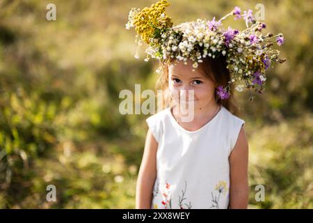 Una giovane ragazza con una corona floreale sorride dolcemente mentre si trova in un prato di fiori selvatici illuminato dal sole, catturando l'essenza dell'innocenza, della natura e dell'infanzia Foto Stock